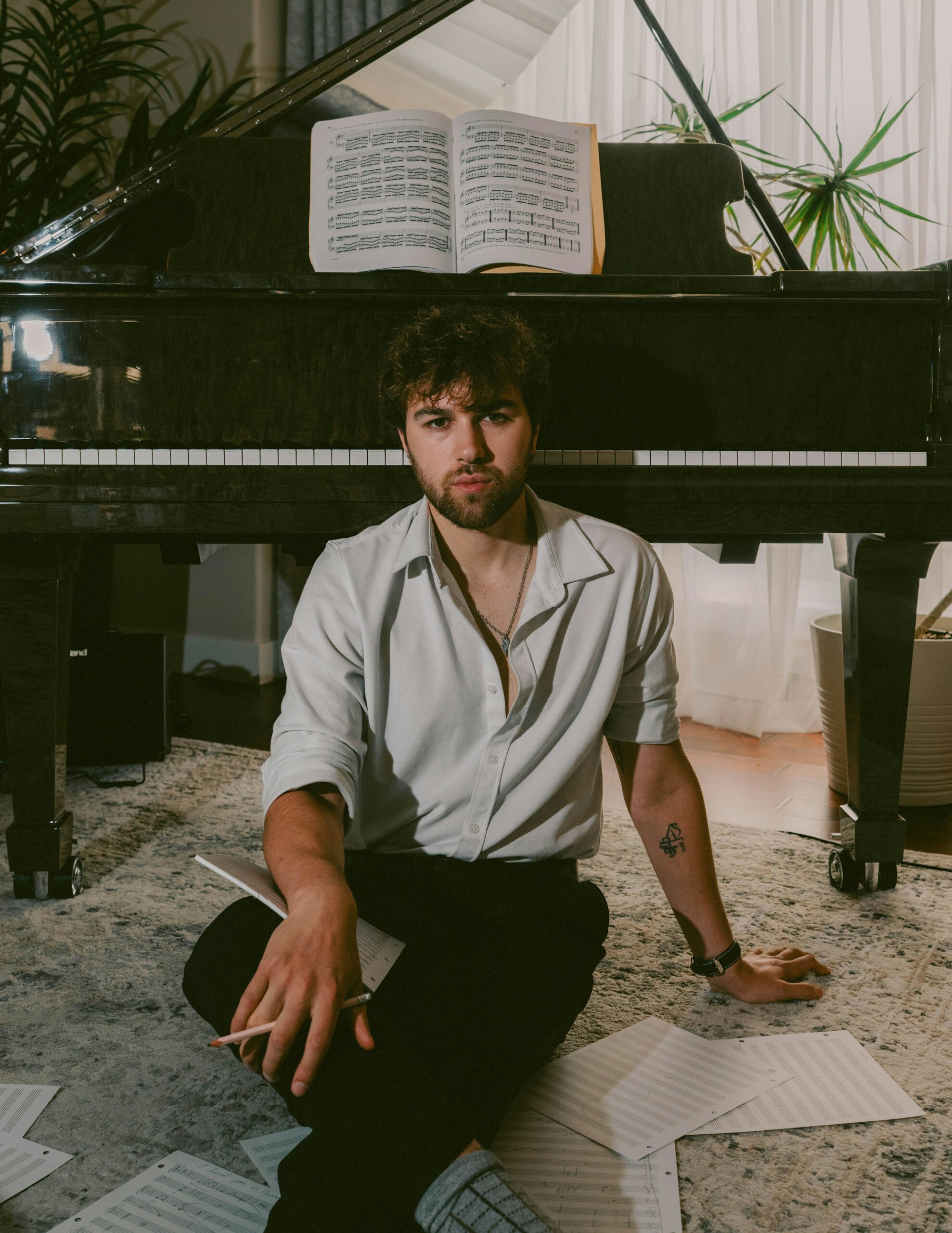 Man with beard sits on a rug in front of a black grand piano, surrounded by scattered sheet music and a pencil in hand.