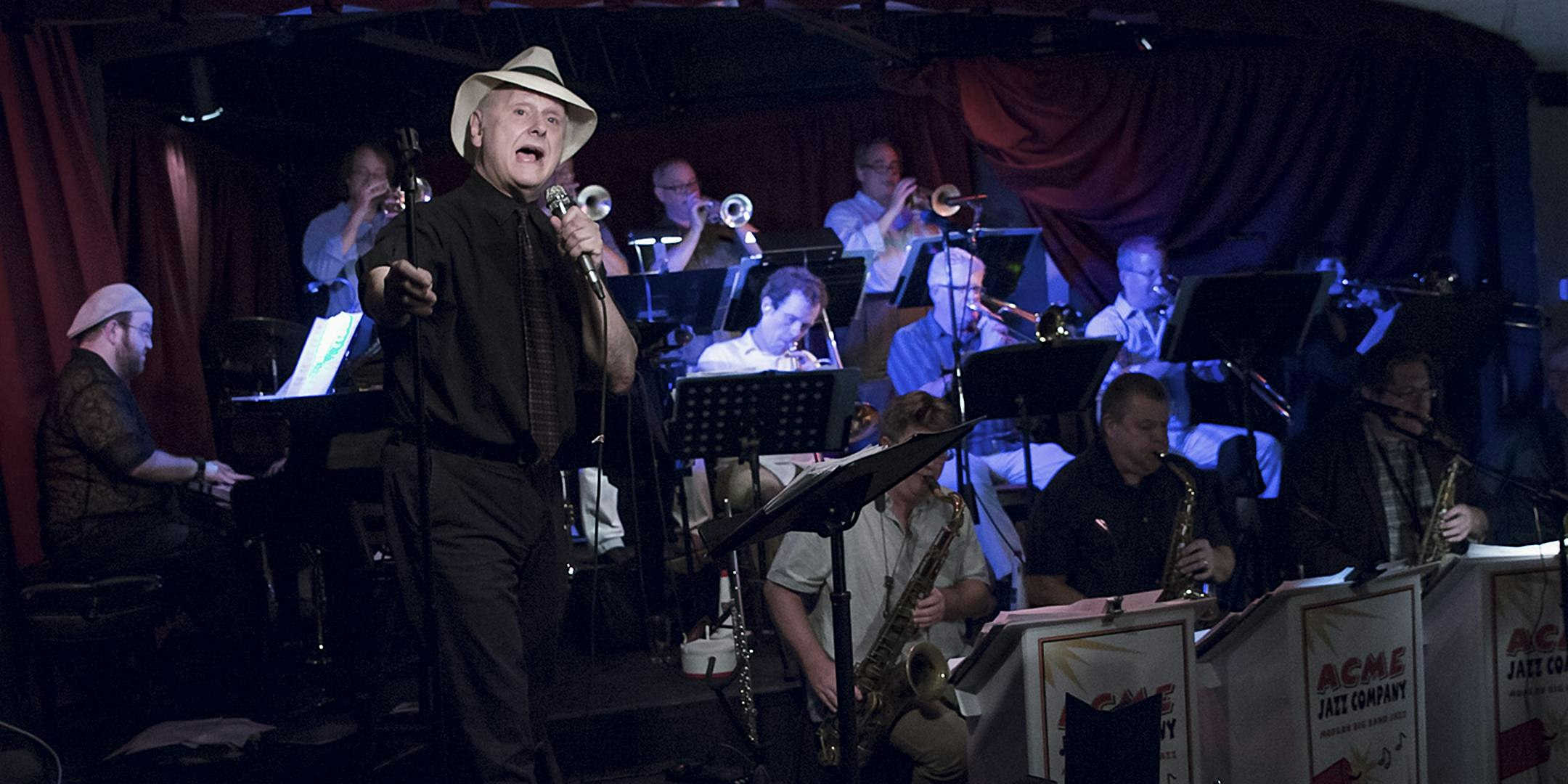 Jazz vocalist in a light fedora sings into a microphone on stage, backed by a large brass and reed ensemble.
