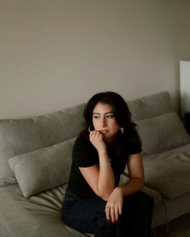 Woman sitting on a gray couch, looking to the side with a thoughtful expression, resting her chin on her hand, wearing a black top and gold jewelry.