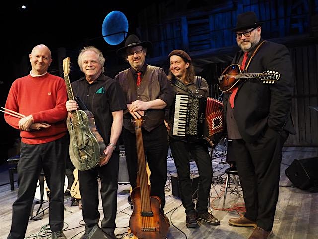 Five male musicians on a stage posing with their instruments: a drummer, resonator guitar, upright bass, accordion, and guitar, wearing casual/retro attire.