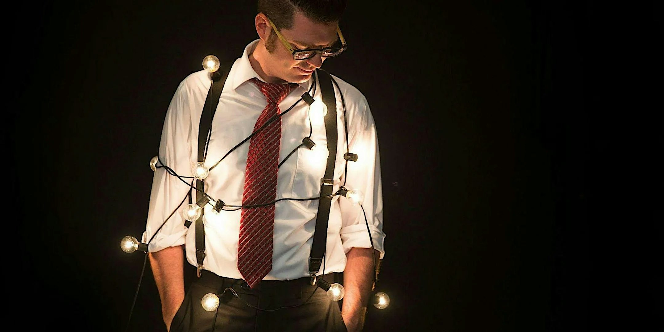 Man in a white shirt and red tie with string lights wrapped around his torso against a dark background.