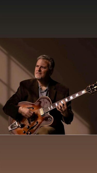 Older man in a dark blazer playing a sunburst hollow-body guitar, looking off to the side as if performing.
