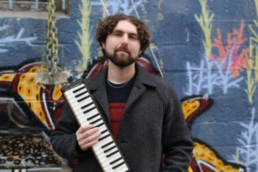 Man with curly hair and beard wearing a dark coat, holding a melodion in front of a colorful graffiti wall.