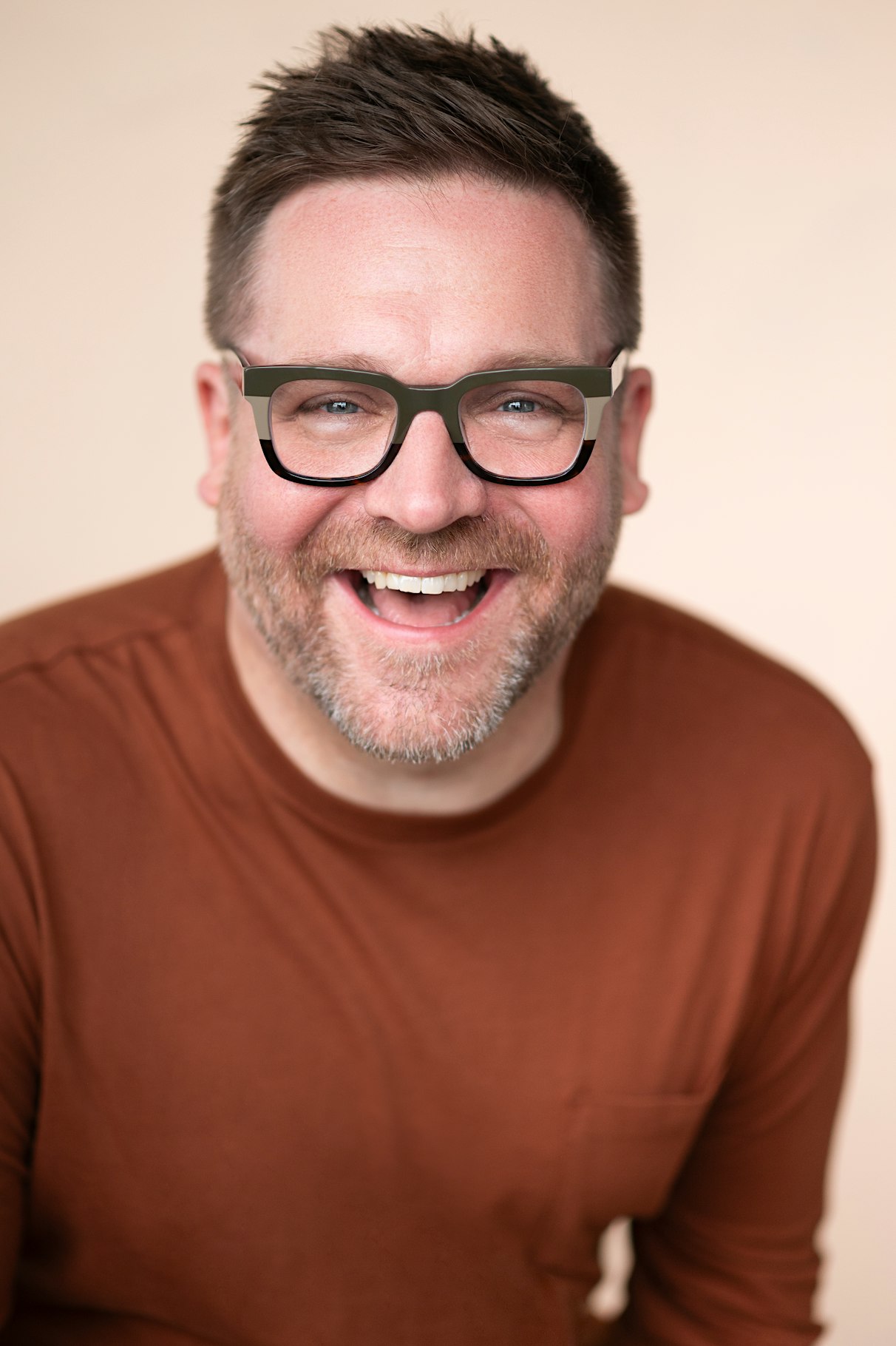 Smiling man with short dark hair and olive-green glasses wearing a brown shirt against a beige background.